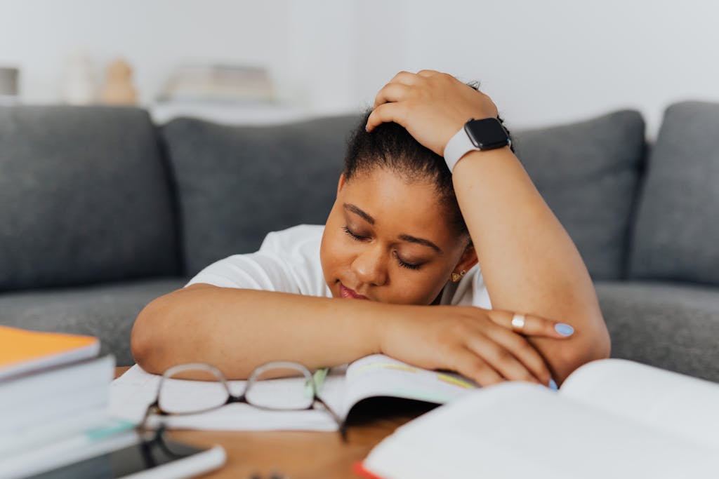Black woman leaning on books with eyes closed, looking tired and distracted while studying on the couch.