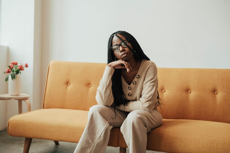 Thoughtful young African American female in casual outfit with eyeglasses looking at camera while sitting on couch with hand on face near table with flowers in vase in bright house