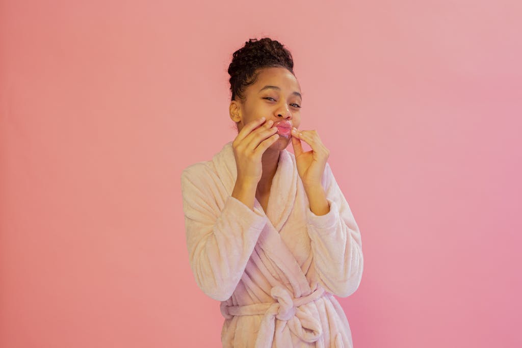 Young African American woman in a pink bathrobe applying a skincare sheet mask against a pink background.